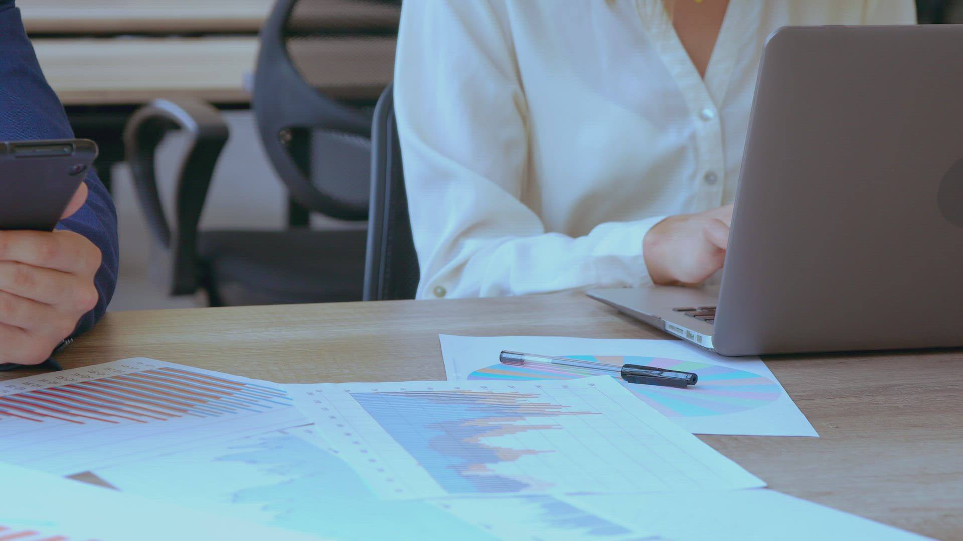 Two colleagues at a desk: one checks a phone while the other types on a laptop beside printed charts.
