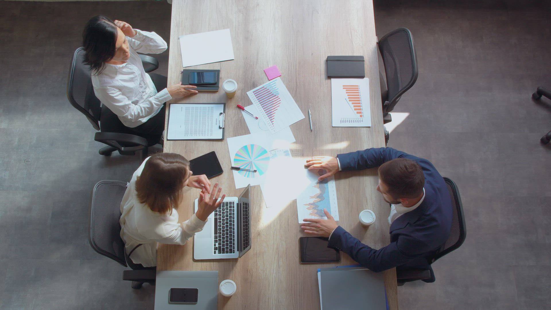 Colleagues around a table of charts and documents, seen from above.