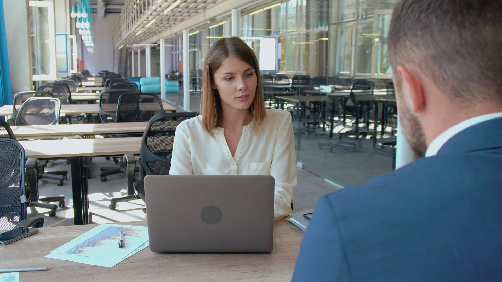 A woman works at a laptop across the desk from a colleague.