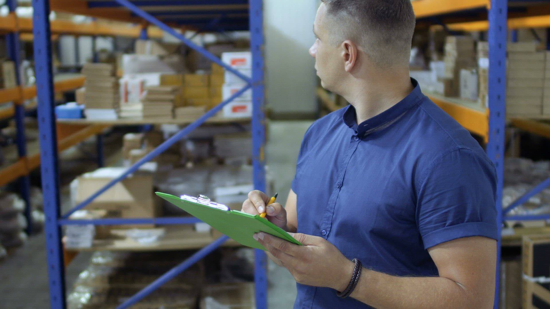 A warehouse worker checks stock on a clipboard between shelves.